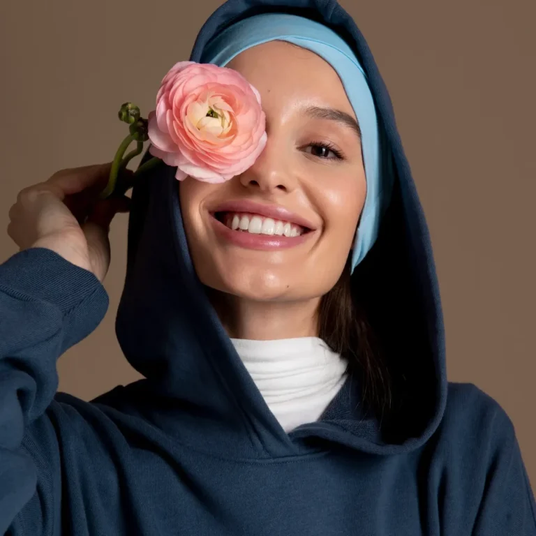Femme souriante en sweat à capuche, tenant une fleur rose près du visage.