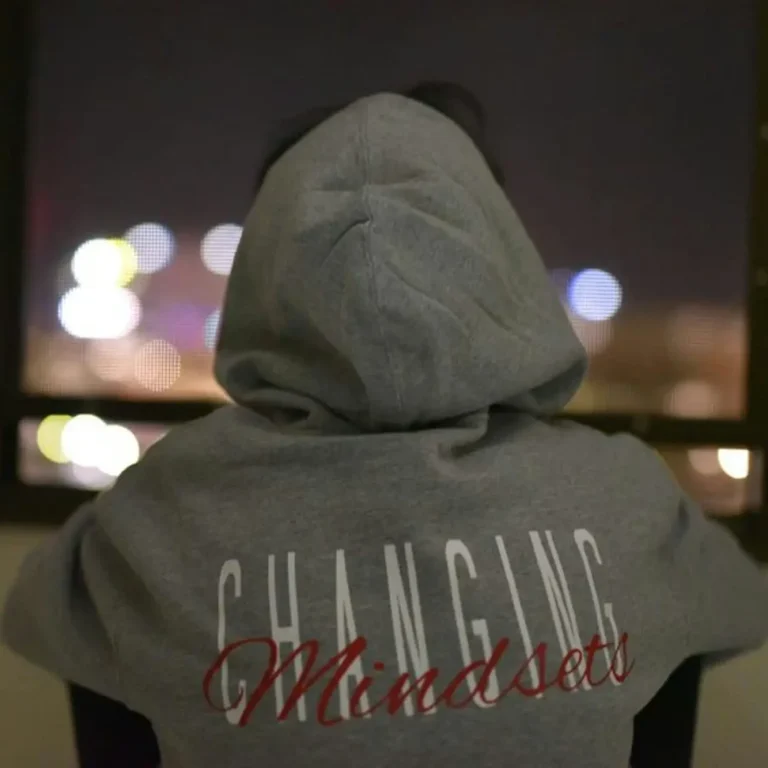 Personne regardant à travers une fenêtre, portant un sweat gris avec "Changing Mindset" au dos.