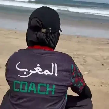 Coach sportive pour femmes Muslima Fit assise sur la plage, portant un vêtement avec écrit "المغرب" et "COACH" au dos.
