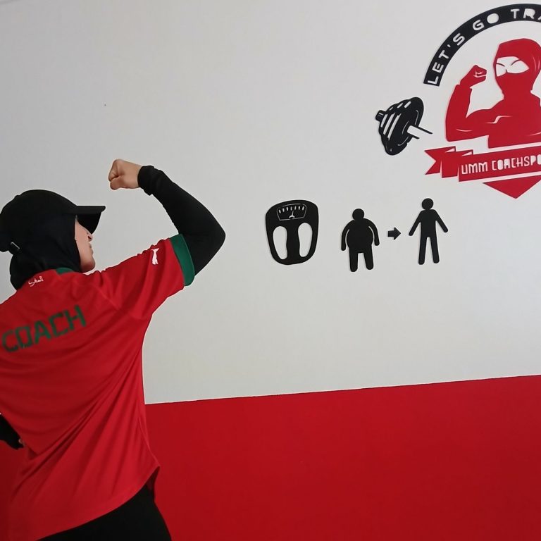 Muslima fit en tenue de coach levant le bras, avec des illustrations murales motivantes.