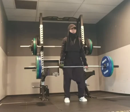 Muslima Fit en tenue de sport soulevant une barre avec des poids dans une salle de musculation.