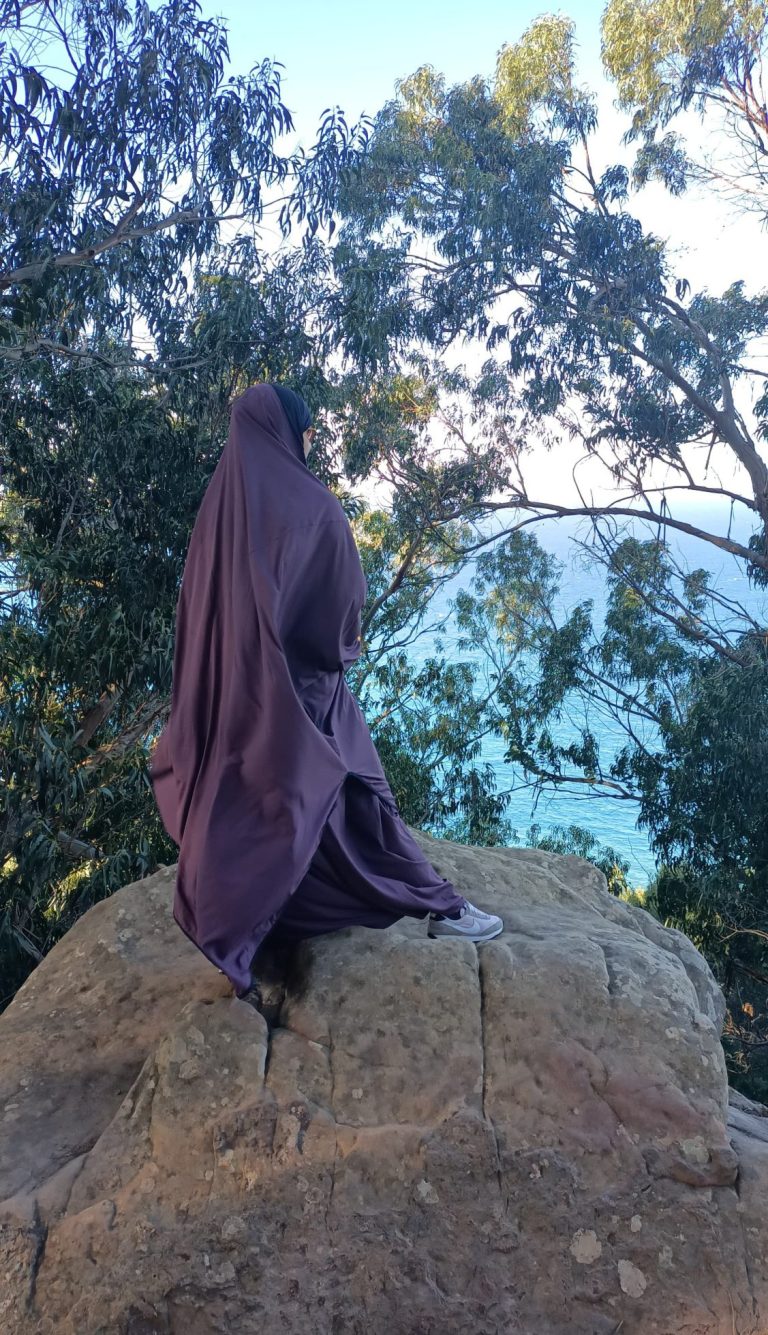 Coach Muslima Fit souriante en hijab, posant près de rochers au bord de la mer.