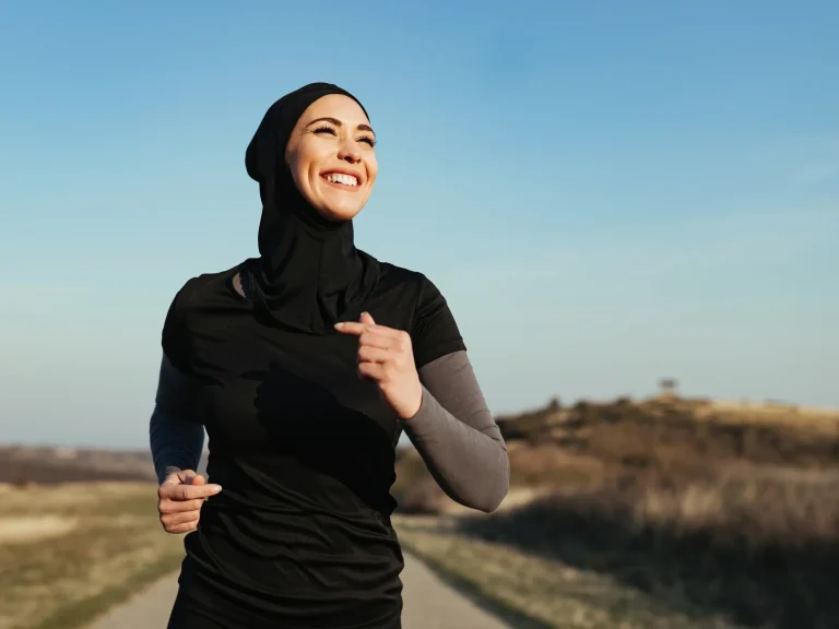 Femme en hijab courant sur un chemin en plein air, souriante sous un ciel bleu en toute liberté.