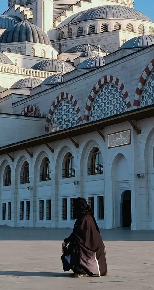 Femme en habit traditionnel musulman, assise devant une mosquée ornée de motifs.