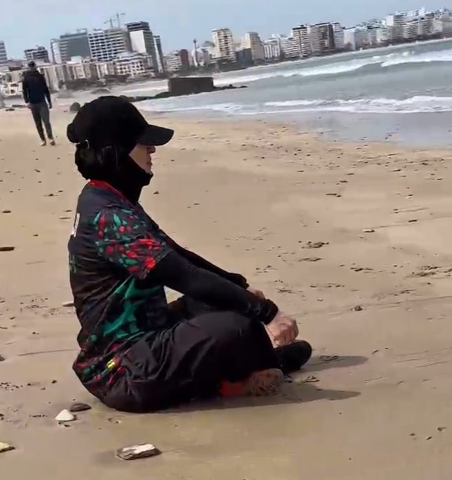 Muslima Fit assise sur la plage, regardant la mer, portant un chapeau et un pull.