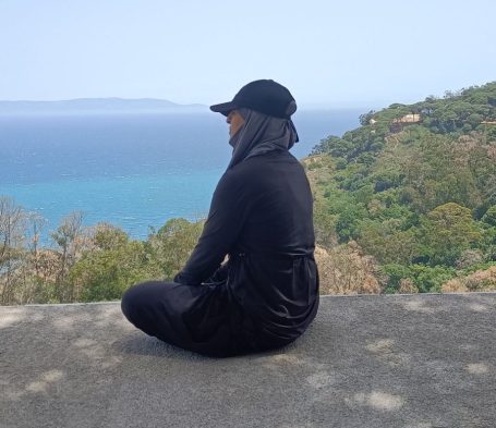 Muslima Fit assise sur un mur surplombant une mer turquoise et une végétation verdoyante.